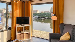 Uma sala de estar moderna com uma TV grande em um suporte branco, um sofá riscado em preto e branco e vista para um canal com um barco atracado.