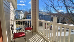 A covered porch with two metal chairs and a red cushion, a white fence, and a view of neighboring houses and trees.