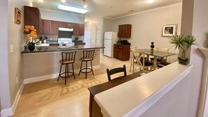 A modern kitchen with a central island, bar stools, a dining table with chairs, and a small dining area with a plant.