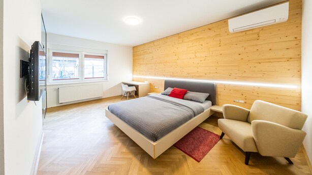 A modern bedroom with a wooden headboard, a bed with grey bedding, a red pillow, a beige armchair, and a TV mounted on the wall. - Apartments Kamr'ca - room 2 & 5 with private bathroom (Ljubljana)