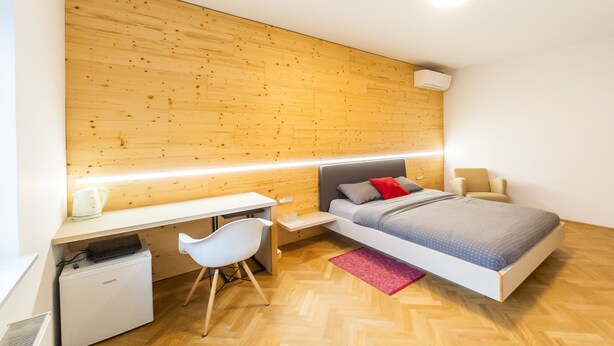 A modern bedroom with a wooden headboard, a bed with grey bedding, a white desk with a chair, and a small white radiator. - Apartments Kamr'ca - room 2 & 5 with private bathroom (Ljubljana)