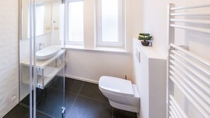 A modern bathroom with a glass shower enclosure, a white sink, a toilet, and a window providing natural light. - Apartments Kamr'ca - room 2 & 5 with private bathroom (Ljubljana)
