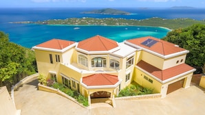 A large, multi-story building with a red-tiled roof, surrounded by lush greenery and overlooking a turquoise sea with clear skies.