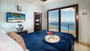 A bedroom with a bed, a nightstand, a painting of a beach, and a view of the ocean through a sliding door.
