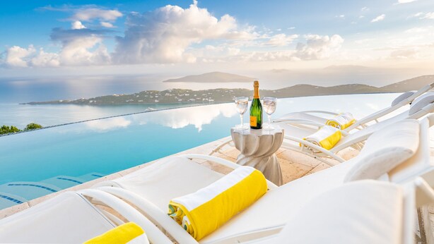 A poolside lounge area with white lounge chairs, a table with a bottle of champagne, and a view of the sea and distant hills.