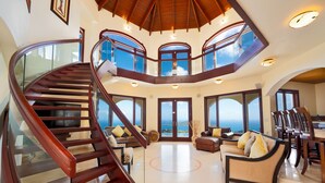 A spacious interior with a grand staircase, multiple seating areas, and large windows offering ocean views.