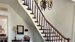 Una scala con balaustre in legno e un lampadario, un tappeto rosso con motivi decorativi, e un muro con un cartello che recita 'LAUGH OFTEN'.