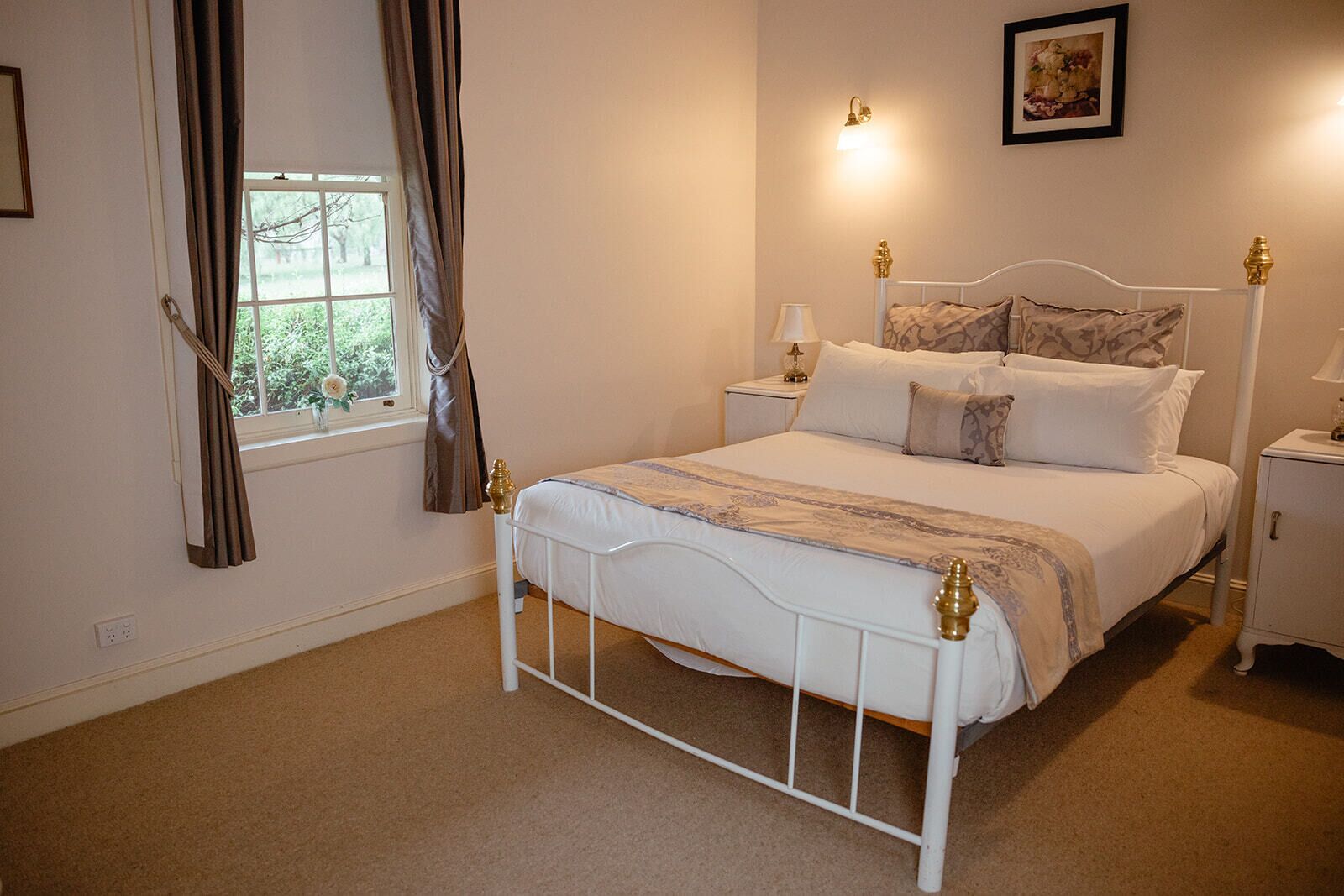 Een slaapkamer met een bed met wit metalen frame, een nachtkastje met lamp en een raam met gordijnen.