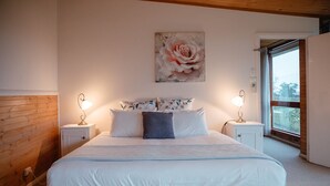 A bedroom with a large bed, two bedside tables with lamps, a floral artwork above the bed, and a window with a view.