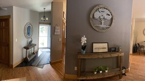 A hallway with a wooden floor, a mirror, a chandelier, a framed picture, a small shelf with plants, and a decorative tree wall hanging. - WolfDen Getaway-Winnebago Channel (Oshkosh)