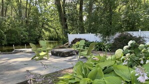 A patio area with green Adirondack chairs, a stone fire pit, and lush greenery including hostas and hydrangeas. - WolfDen Getaway-Winnebago Channel (Oshkosh)