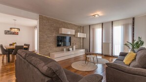 Ein modernes Wohnzimmer mit einer Wand aus Naturstein, einem Flachbildfernseher, einem Essbereich mit Tisch und Stühlen und einer gemütlichen Sitzecke mit Sofas.