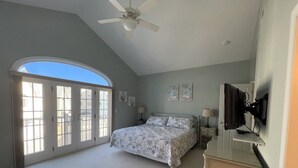 A bedroom with a bed, a dresser, a TV, and a ceiling fan. There are windows and a door leading outside.