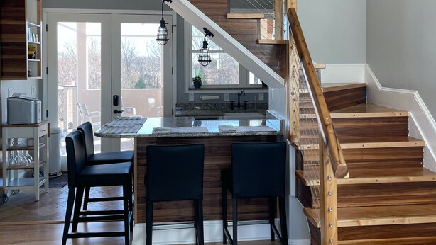 A modern kitchen with a wooden staircase, a dining area with bar stools, and a view of the outdoors.