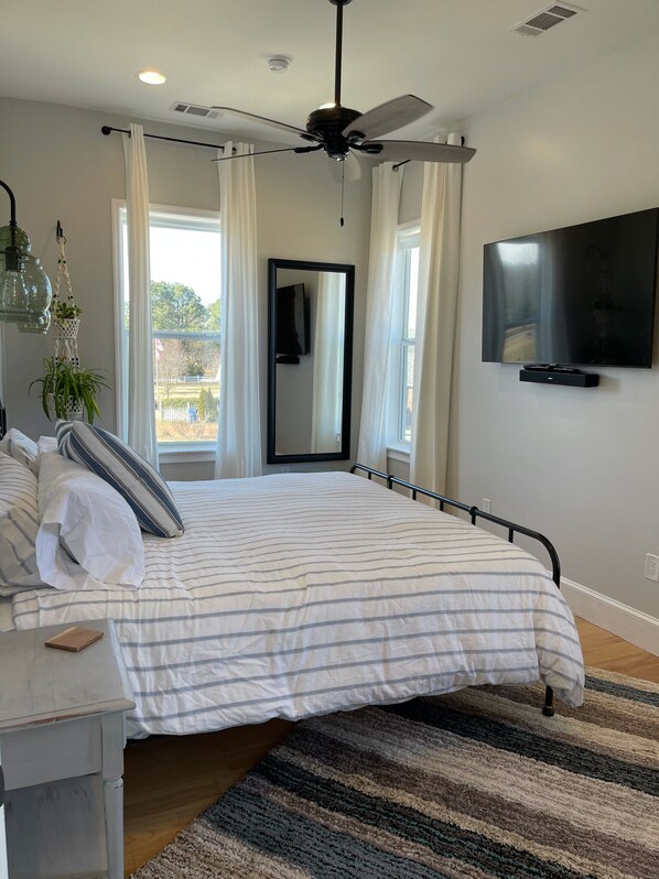 A bedroom with a bed, bedside table, mirror, ceiling fan, TV, and large windows with curtains. - Memphis Meadows - Collierville Home - privacy with great views (Collierville)