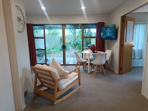 A room with a wooden sofa, a dining table with chairs, a TV, and a sliding glass door leading to a garden. - Beach Bach (The Pines Beach)