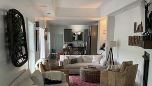 A spacious living room with a white sofa, a beige sofa, and a glass coffee table. There is a red rug on the floor and a kitchen in the background. - Lake Rabun Rental - Classic Waterside Home and Boathouse (Lakemont)