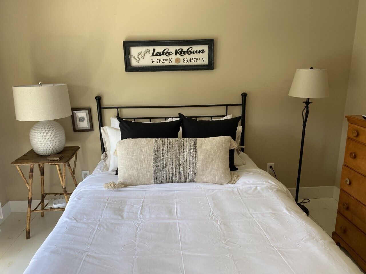 A neatly made bed with white bedding, a black metal headboard, and a framed sign above it.