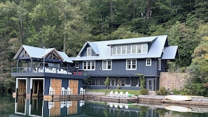 A large, multi-story house with a dock extending into a lake, surrounded by dense forest. - Lake Rabun Rental - Classic Waterside Home and Boathouse (Lakemont)