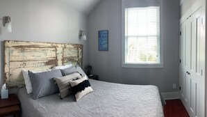 A bedroom with a large bed, a rustic headboard, a window with blinds, and a door with a framed picture.