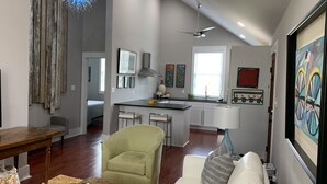 A spacious living room with a white sofa, a green armchair, and a wooden coffee table. A kitchen with a central island and a ceiling fan is visible in the background.