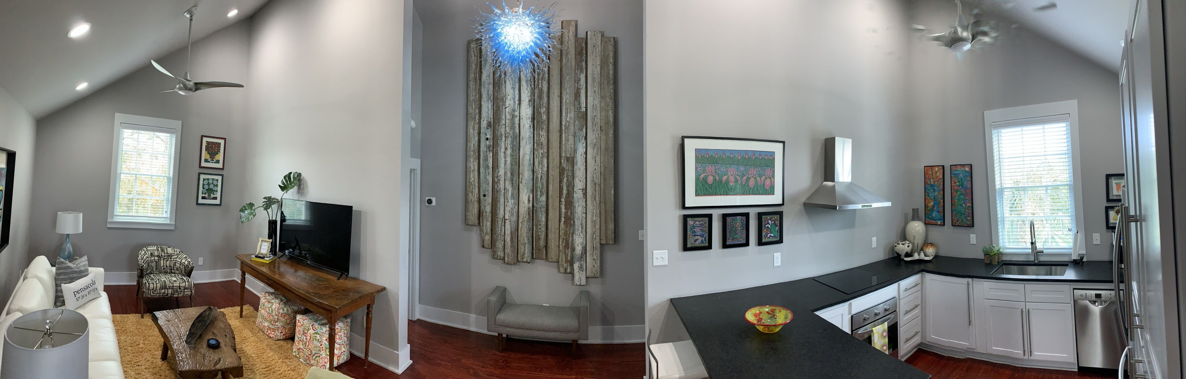 Amplia sala de estar con un televisor de pantalla plana grande, una mesa de centro de madera y un sillón estampado. La cocina cuenta con gabinetes blancos, una encimera negra y electrodomésticos de acero inoxidable.