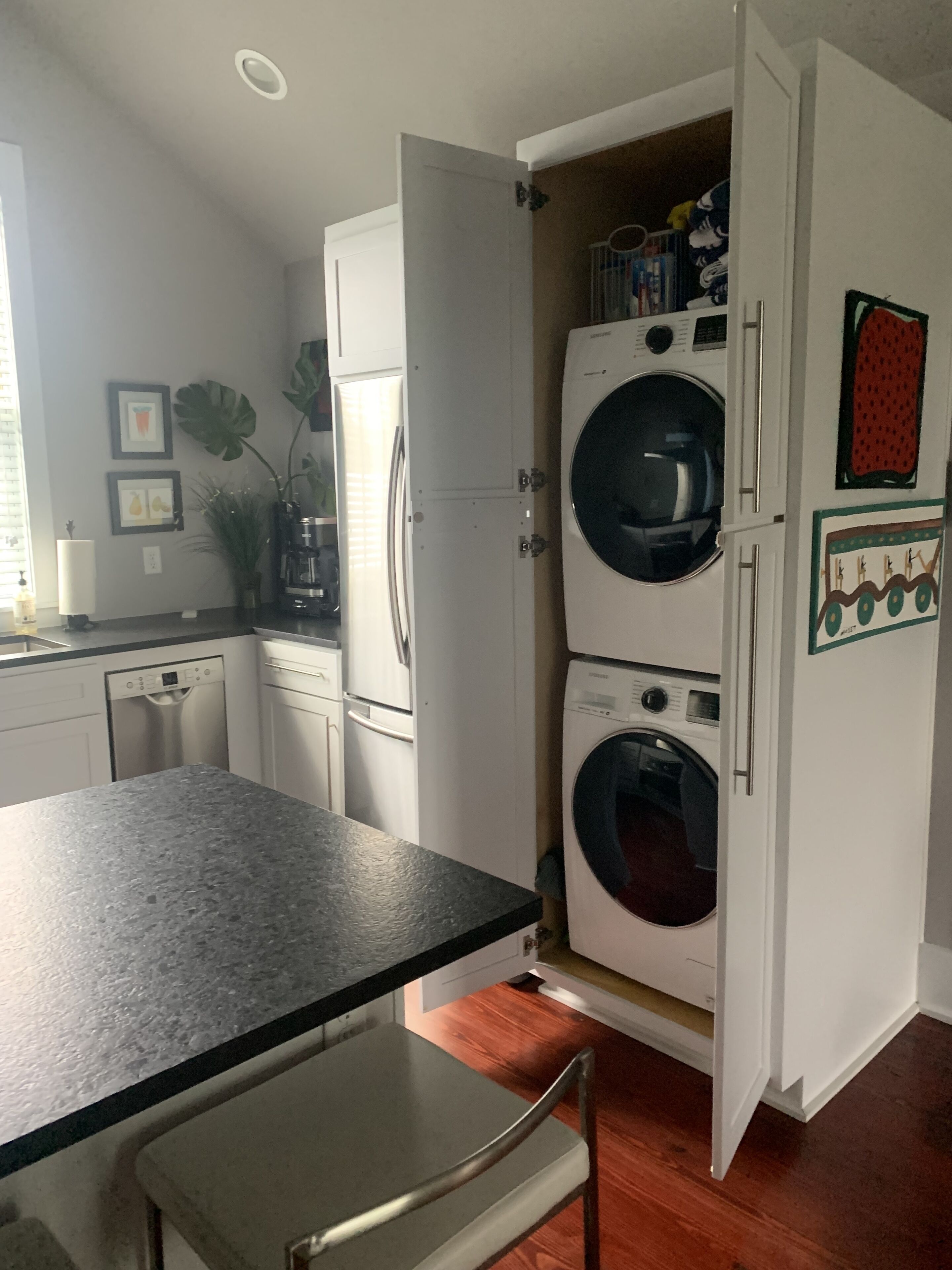 A compact kitchen with white cabinetry, a dishwasher, a double-door washer-dryer unit, and a countertop with a chair.