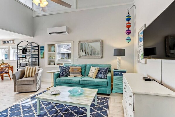 A spacious living room with a teal sofa, a coffee table, and a TV. There is a bookshelf, a chair, and a decorative wall hanging.