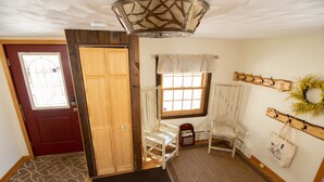 Une pièce avec une porte en bois, une fenêtre avec un rideau, un banc, un porte-manteau orné d’une guirlande, et une petite table sur laquelle se trouve une boîte.