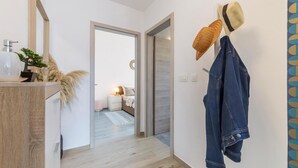 Een hal met een houten vloer, een witte muur en een deur naar een slaapkamer. Er staat een kapstok met een hoed en jas.