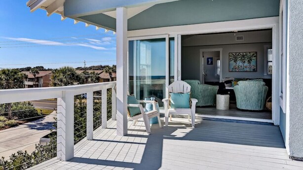 Un balcón con barandilla blanca, una puerta de vidrio que da a una habitación con un sofá verde y un cuadro, y vistas del entorno.