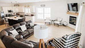 A modern living room with a brown leather sofa, a white table, and a striped armchair. There is a kitchen in the background with white cabinets and a TV mounted above the fireplace.