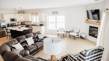 Un salon moderne avec un canapé en cuir marron, une table blanche et un fauteuil à rayures. Au fond, on aperçoit une cuisine avec des meubles blancs et un téléviseur fixé au-dessus de la cheminée.