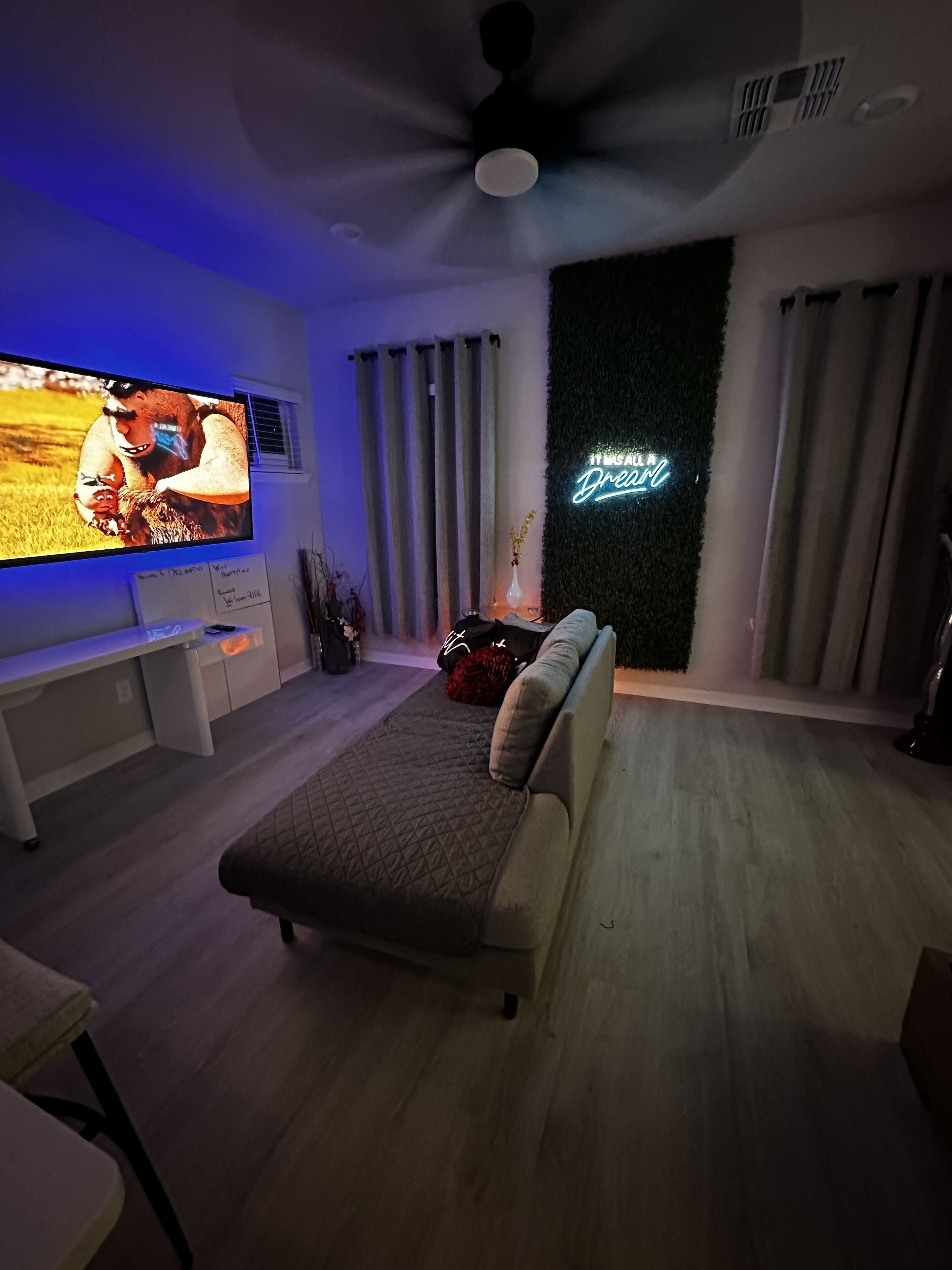 A modern living room with a flat-screen TV, a bed, a neon sign, and a ceiling fan.