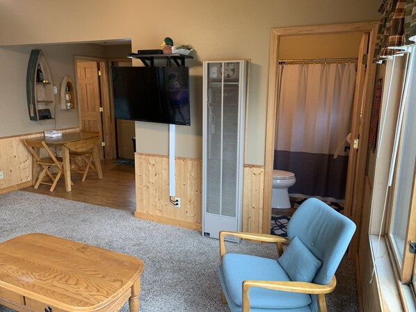 Un espacio habitable pequeño con una mesa de comedor de madera, un sillón azul, un televisor montado en un mueble bajo, un inodoro blanco y una zona de ducha.