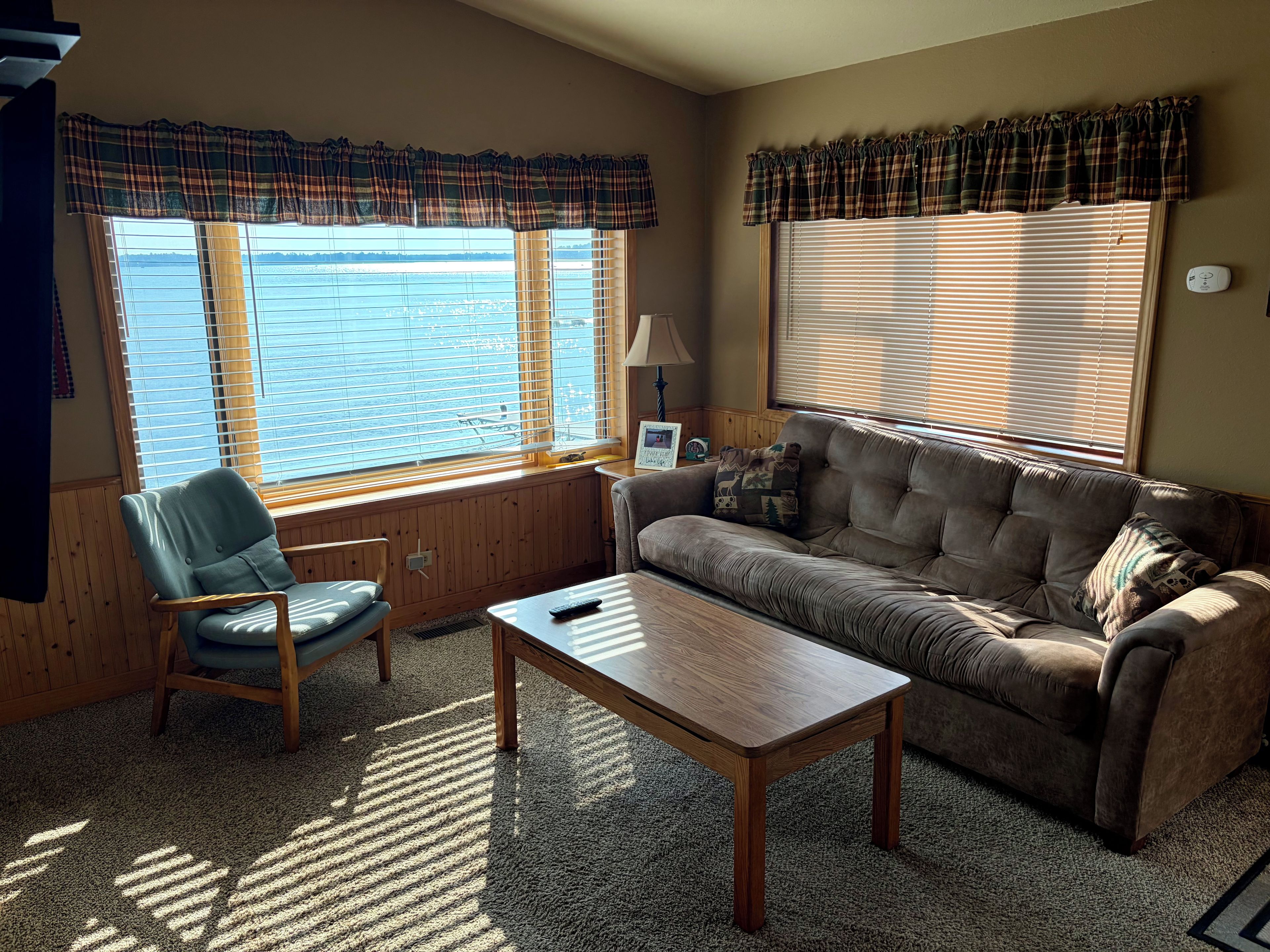 A furnished living room with a sofa, armchair, and coffee table, large windows with blinds, and a view of the outdoors.