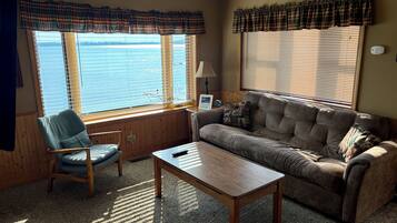 A furnished living room with a sofa, armchair, and coffee table, large windows with blinds, and a view of the outdoors.