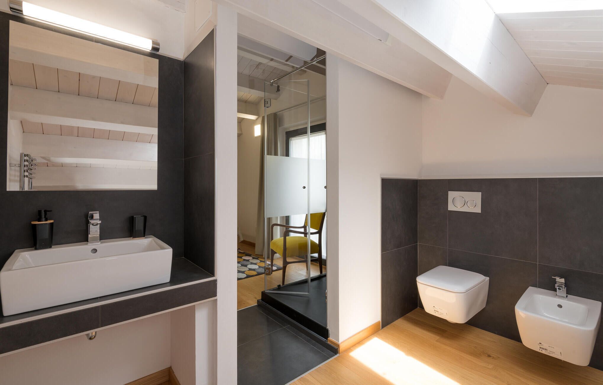 Un baño moderno con un espejo grande, un lavamanos blanco y un área de aseo separada.