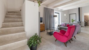 A modern living room with a staircase, a grey sofa set, a vibrant pink chair, and a flat-screen TV.