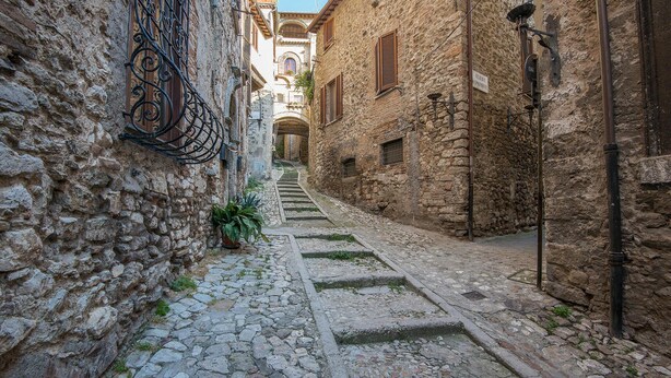 A narrow cobblestone street flanked by historic stone buildings with wooden shutters and iron railings.