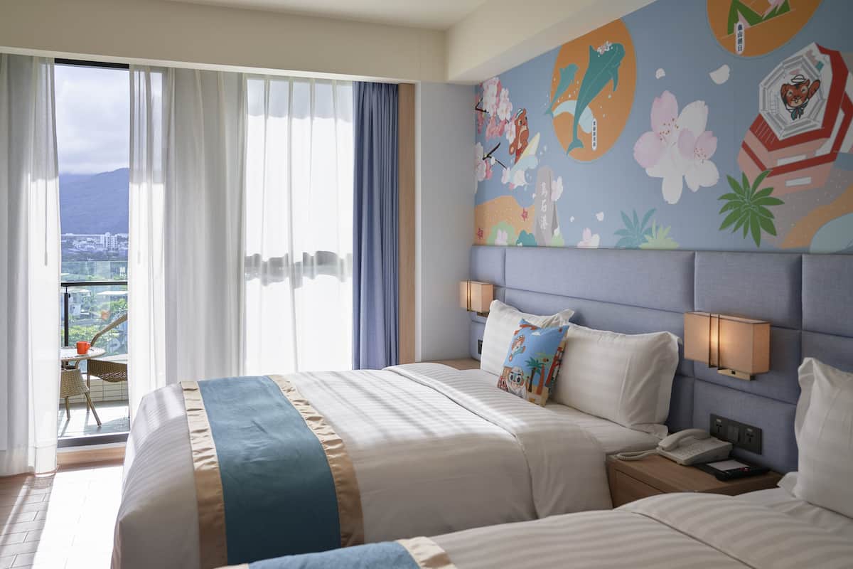 A hotel room with a large bed, a nightstand, a wall-mounted lamp, and a view of a cityscape through a sliding door.