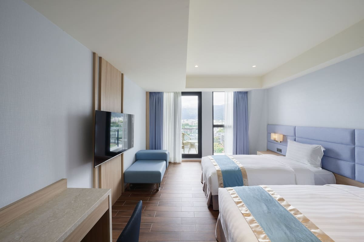 A hotel room with two beds, a TV, a desk, and a view of the city through large windows.