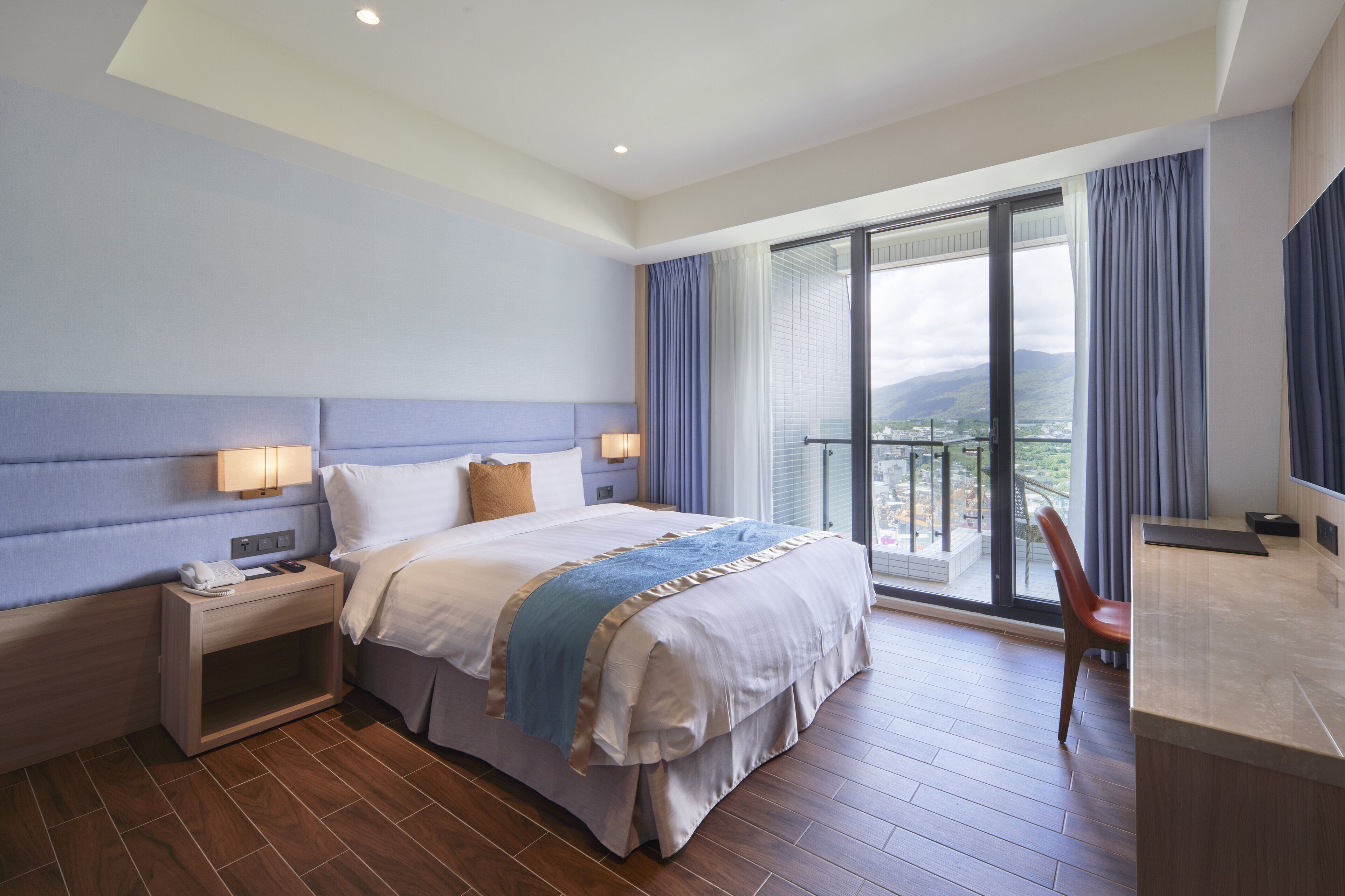 A hotel room with a large bed, a desk, a chair, and a balcony with a view.