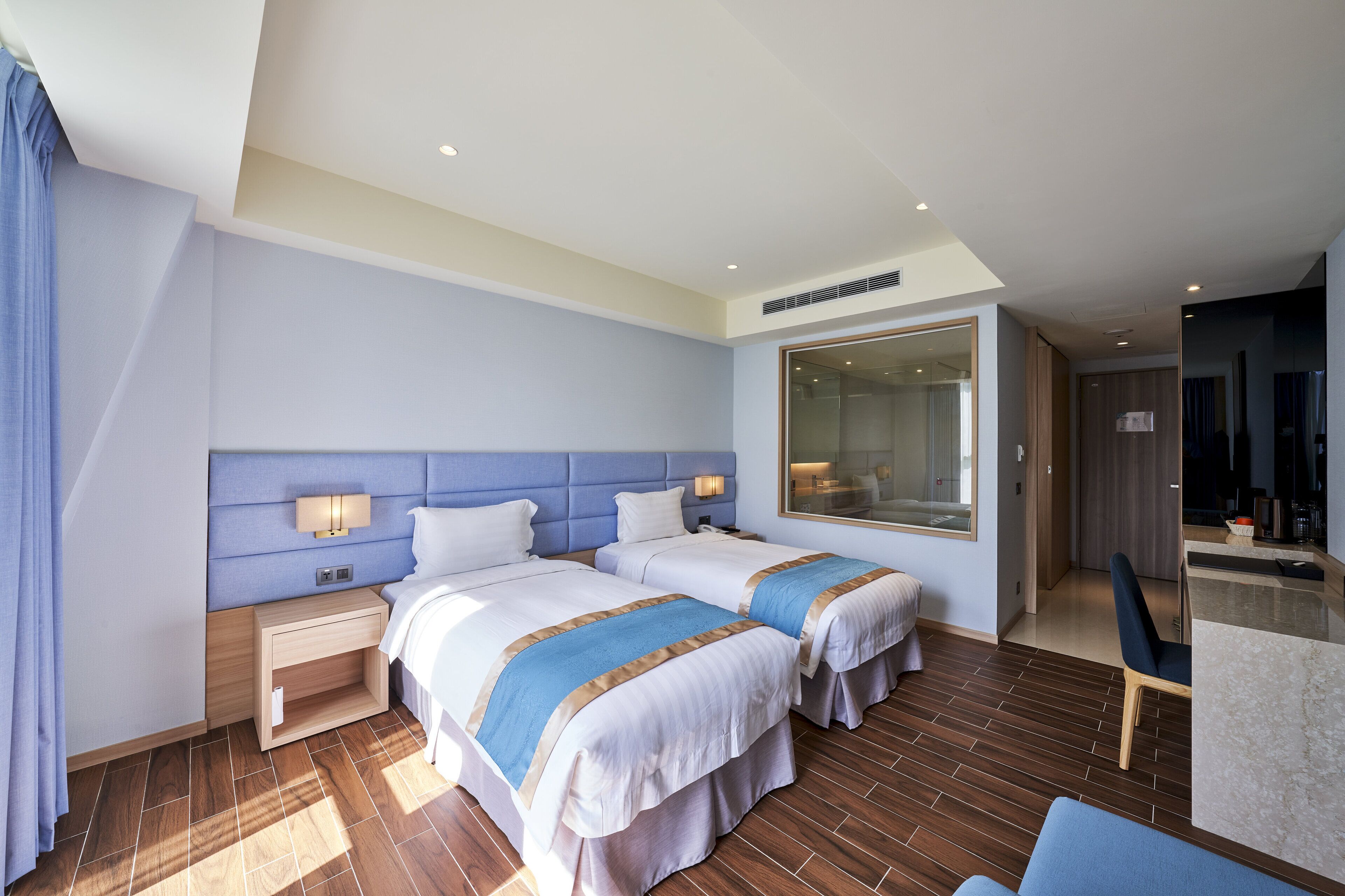 Ein Hotelzimmer mit zwei Betten, einem großen Spiegel, einem hölzernen Nachttisch und einem modernen Badezimmer, das durch eine geöffnete Tür sichtbar ist.