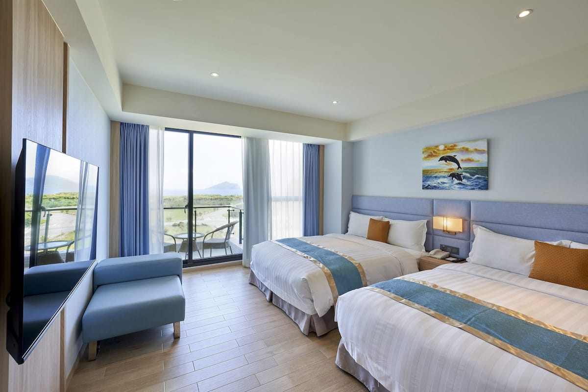 A hotel room with two beds, a TV, a balcony with a view, and a painting on the wall.