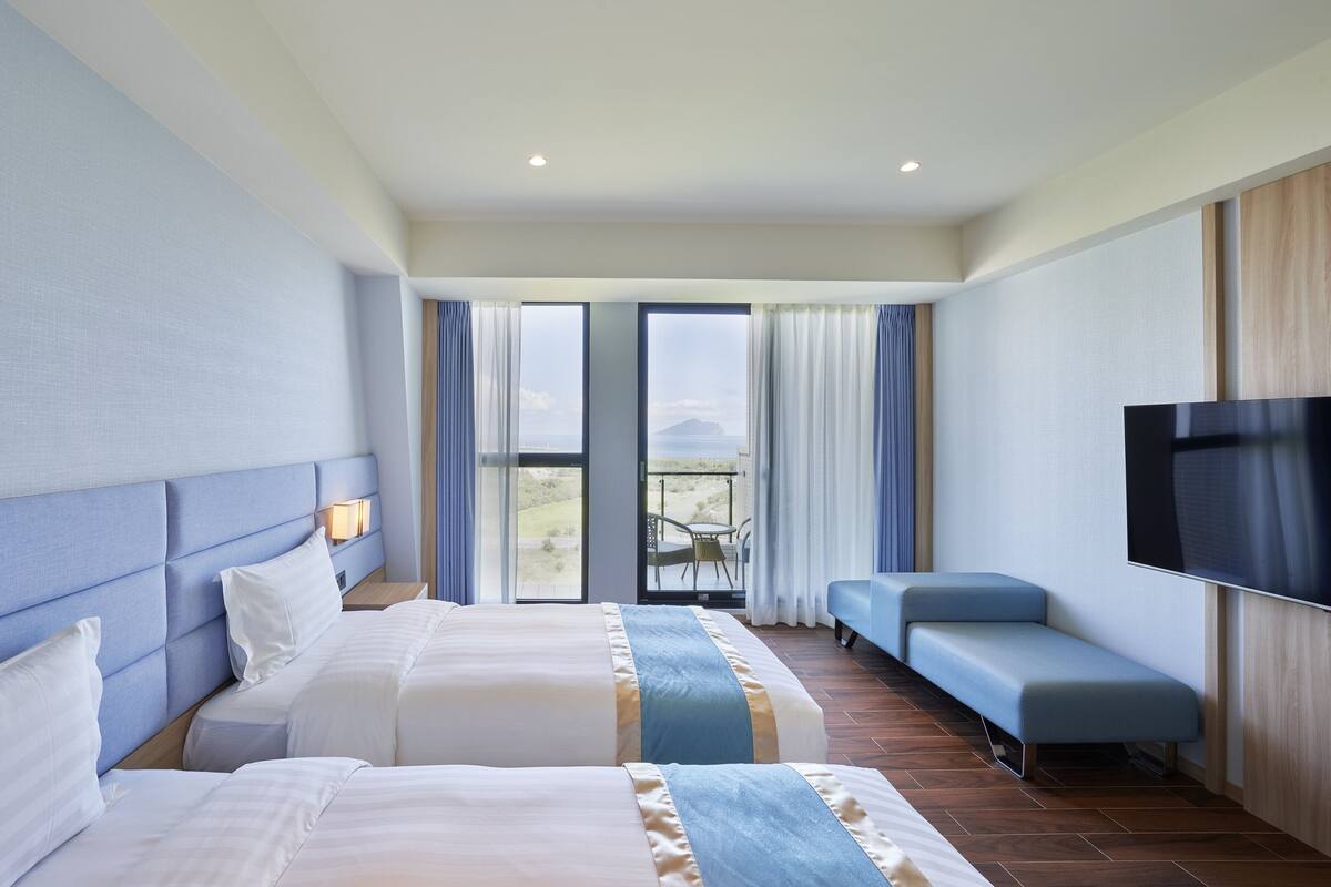 A hotel room with two beds, a TV, a balcony with a view, and a seating area.