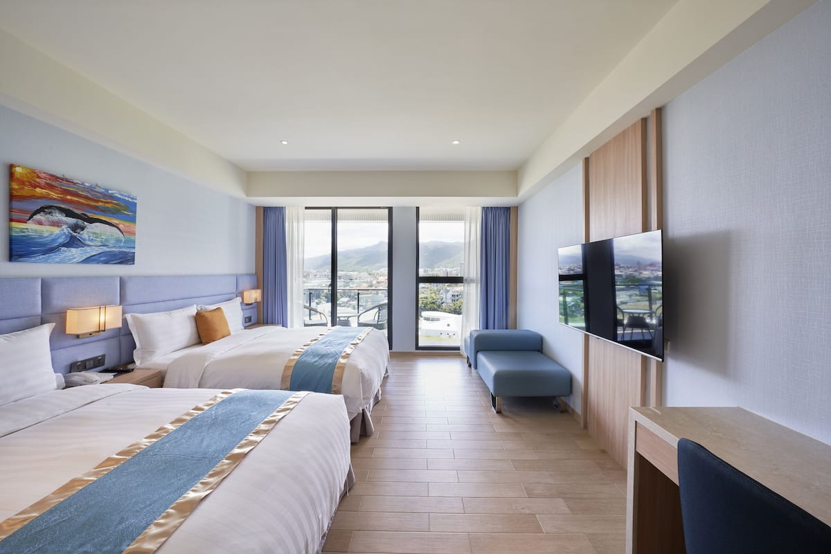 A hotel room with two beds, a TV, a desk, and a view of the city.