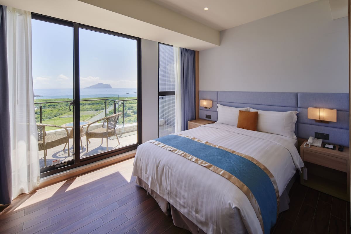 A hotel room with a large bed, a nightstand with a phone, and a view of the ocean through a sliding door.