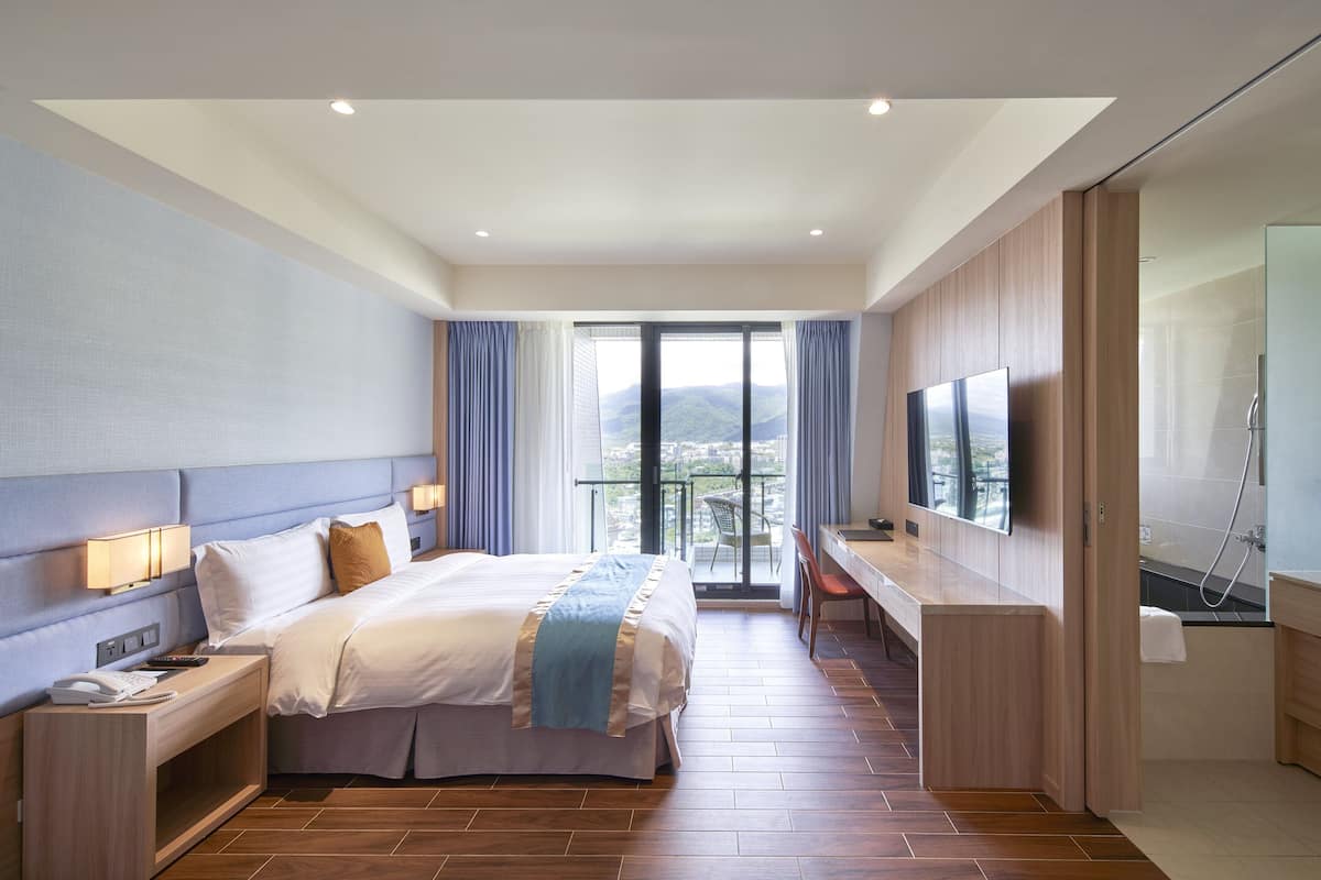 A modern hotel room with a large bed, a desk, a TV, and a balcony with a view.