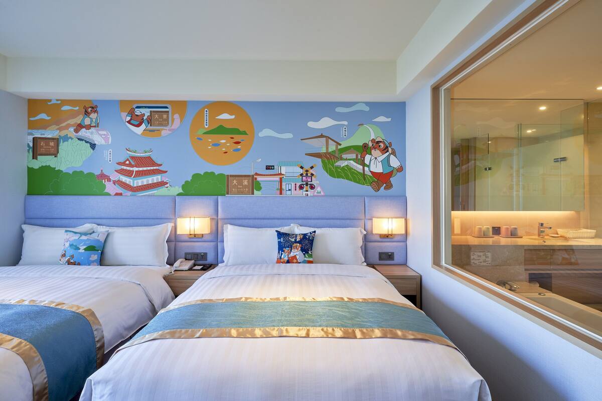 A hotel room with two beds, a large wall mural, and a view of the city through a large window.
